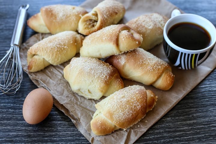 Freshly baked Pan de Coco rolls sprinkled with sugar, arranged on a brown parchment paper with a whisk, a whole egg, and a cup of coffee on the side.