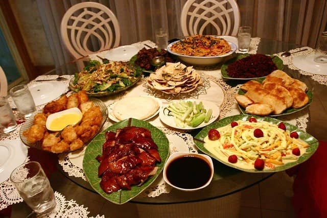 The image shows a festive Christmas feast (Panghanda sa Pasko) with a variety of Filipino dishes displayed on a table. The spread includes fried chicken, grilled meats, fresh salads, pasta, and other traditional Filipino holiday delicacies, all arranged beautifully for a family celebration.