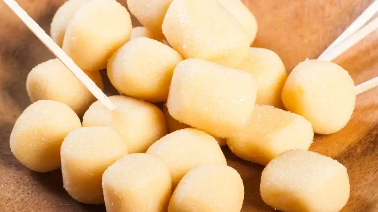 Soft and creamy Pastillas candies, a traditional Filipino milk-based sweet, are neatly arranged on a wooden platter. The small cylindrical treats are dusted with sugar, with toothpicks inserted for easy serving.