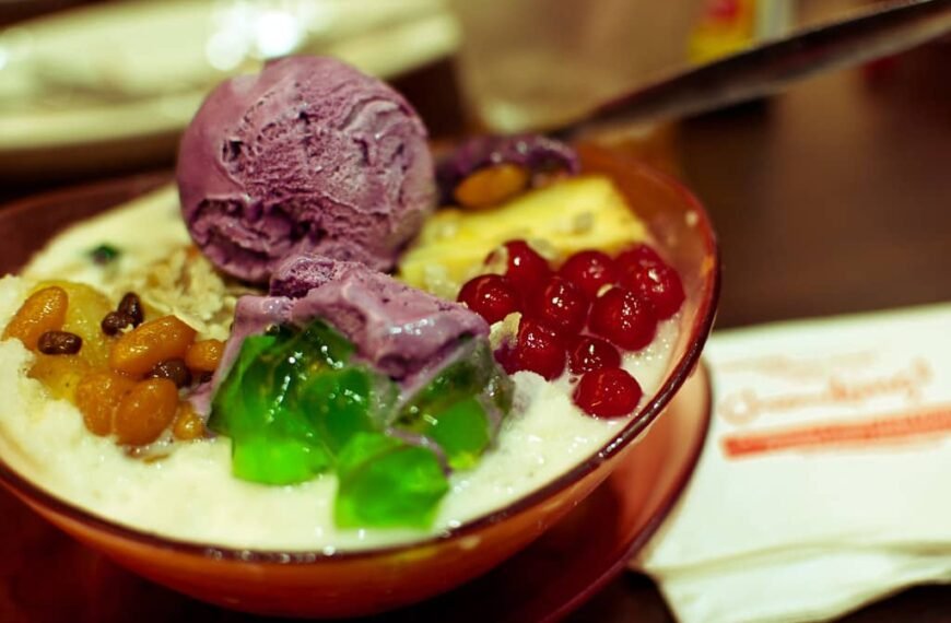 A vibrant bowl of Pinoy Halo-Halo, a popular Filipino dessert, featuring a colorful mix of ingredients. On top, there are scoops of creamy ube (purple yam) ice cream. Surrounding it are sweetened red beans, green gelatin cubes, red tapioca pearls, and candied fruits.