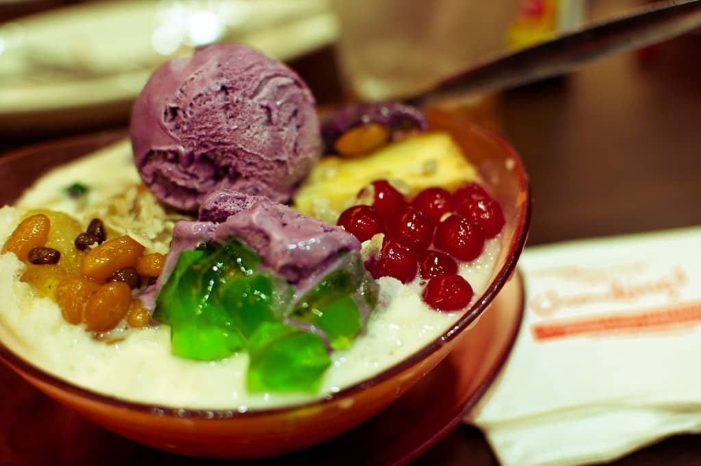 A vibrant bowl of Pinoy Halo-Halo, a popular Filipino dessert, featuring a colorful mix of ingredients. On top, there are scoops of creamy ube (purple yam) ice cream. Surrounding it are sweetened red beans, green gelatin cubes, red tapioca pearls, and candied fruits.