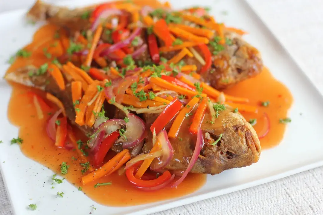 A plate of Pinoy-style Escabeche, a Filipino dish of fried fish topped with a colorful mix of vegetables like carrots, red bell peppers, and onions, all drizzled with a tangy sweet and sour sauce. The fish is garnished with chopped parsley and served on a white dish.