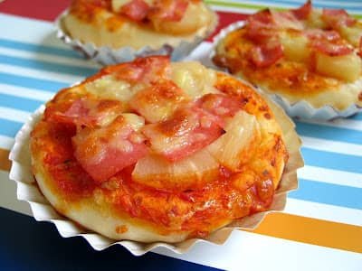 Mini pizza pandesals topped with melted cheese, diced ham, and pineapple chunks, served in small paper cups. These bite-sized Filipino pizzas are laid out on a colorful striped background, combining the fluffy pandesal bread base with savory pizza toppings, creating a unique fusion of flavors.