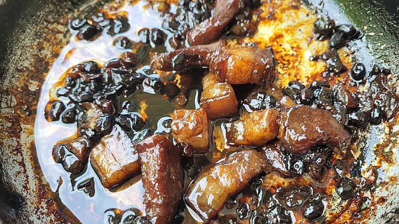 A close-up image of Pork Humba, a Filipino dish made of braised pork belly simmered in soy sauce, vinegar, and black beans, with tender meat and a rich sauce served in a pan.