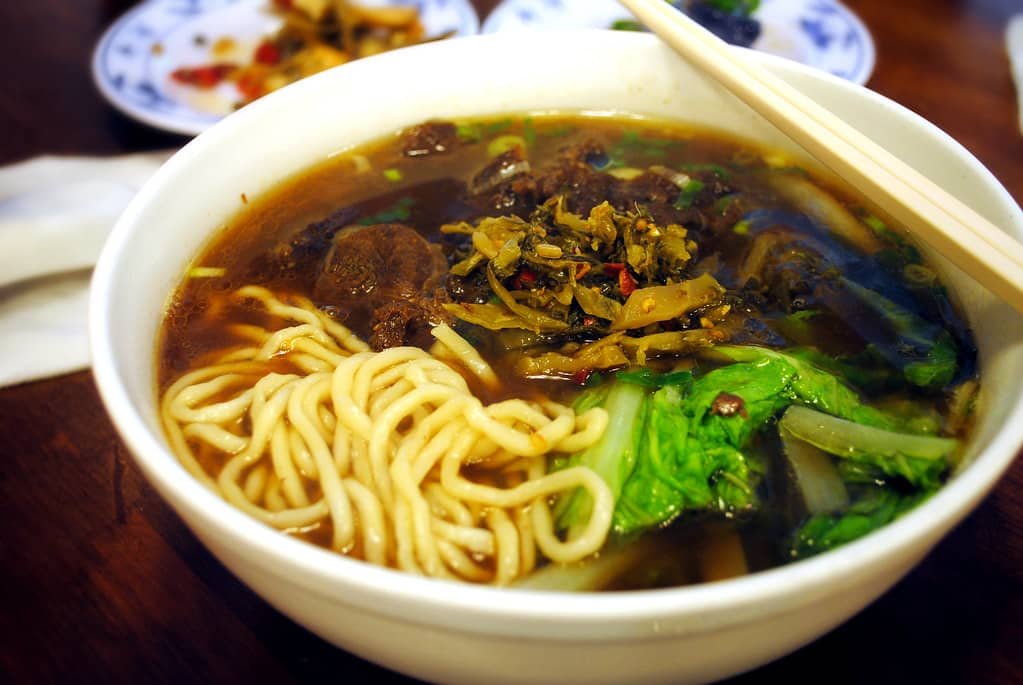 A steaming bowl of Presidential Beef Noodle Soup, featuring tender beef chunks, springy noodles, and vibrant greens. The rich broth is infused with savory flavors and topped with pickled vegetables, creating a hearty and comforting dish. Chopsticks rest on the rim of the bowl, ready for a delicious meal.