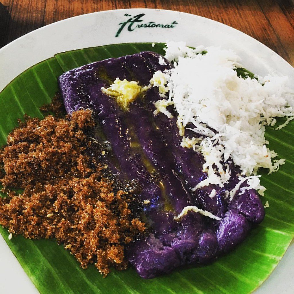 A serving of Puto Bumbong, a traditional Filipino delicacy, placed on a banana leaf. The dish features purple-colored rice cake topped with butter, grated coconut, and muscovado sugar, creating a delightful blend of flavors and textures.