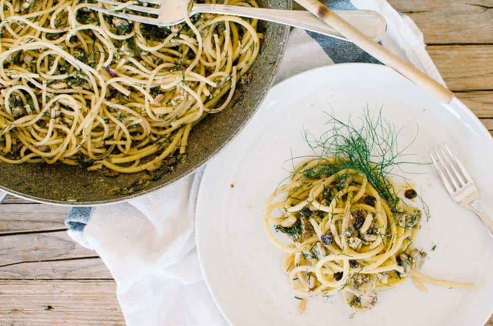 A delicious plate of Sardinas Pasta featuring al dente spaghetti tossed in a savory sardine-based sauce. The dish is garnished with fresh herbs and almond slivers, offering a rustic and flavorful meal. The serving is elegantly presented on a white plate, with additional pasta shown in a pan beside it.