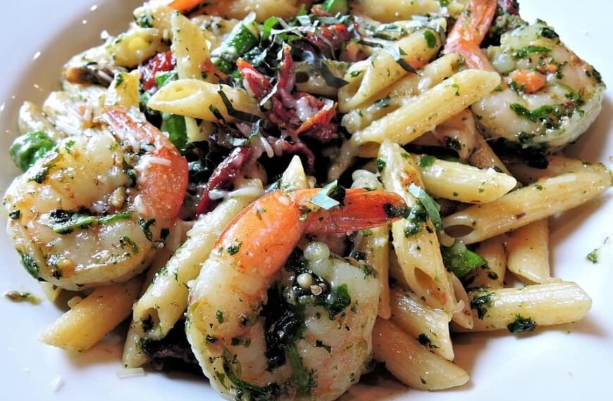 The image shows a plate of Shrimp Mac and Cheese, featuring large shrimp mixed with penne pasta in a creamy cheese sauce, garnished with herbs and spices for added flavor.