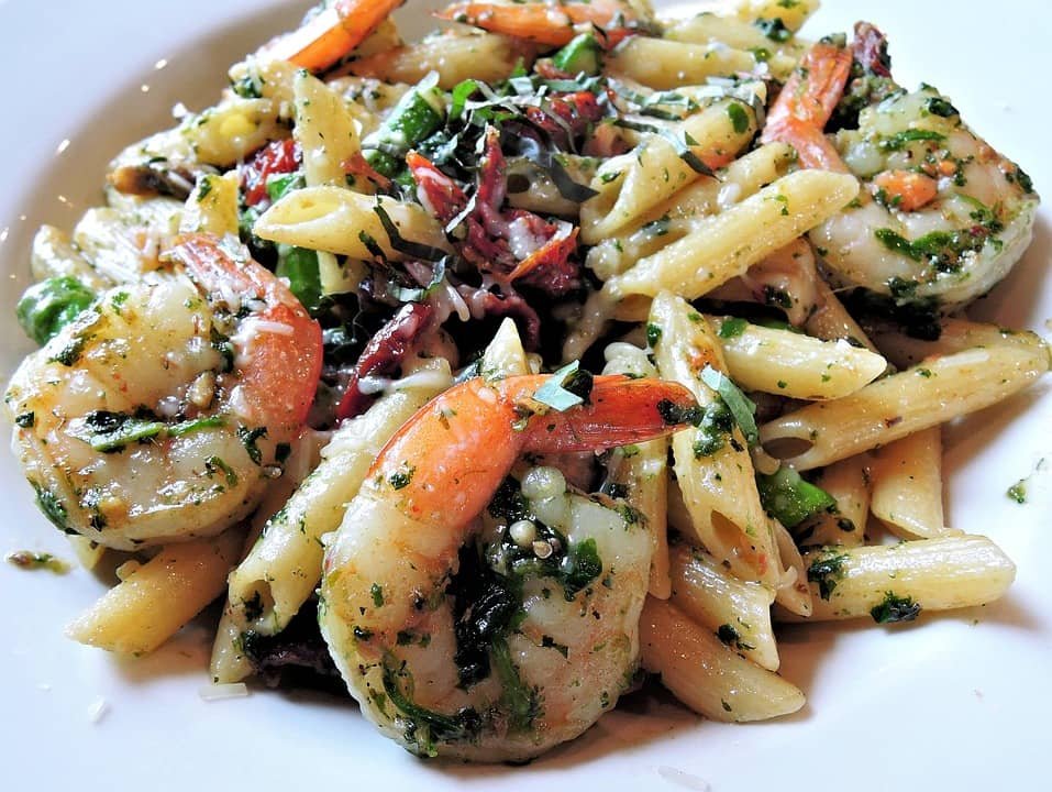 The image shows a plate of Shrimp Mac and Cheese, featuring large shrimp mixed with penne pasta in a creamy cheese sauce, garnished with herbs and spices for added flavor.