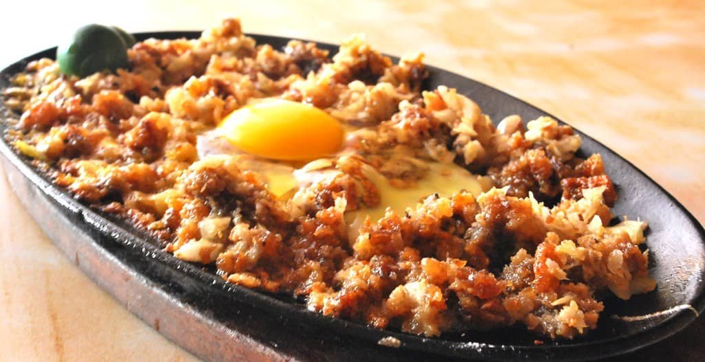 A sizzling plate of Sisig Lutong Bahay, a savory and crispy Filipino dish made from chopped pork, topped with a fresh egg ready to be mixed in, served hot and accompanied by a calamansi for an extra zesty flavor.