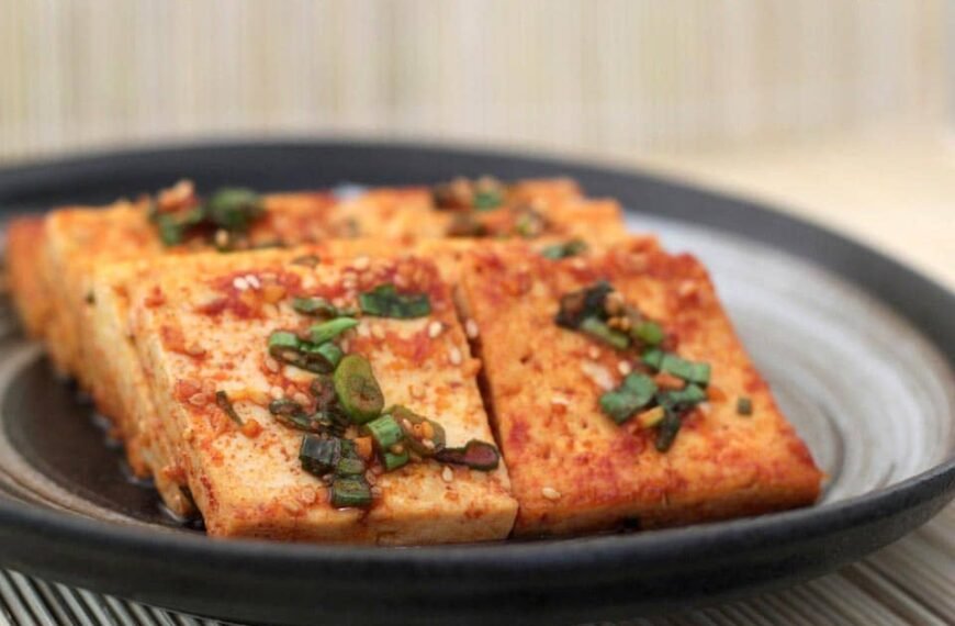 A plate of Sizzling Tofu, featuring golden-brown tofu slices topped with a flavorful sauce, garnished with chopped green onions and sesame seeds. The tofu has a crispy exterior and is seasoned to perfection, offering a delicious and spicy kick in every bite.