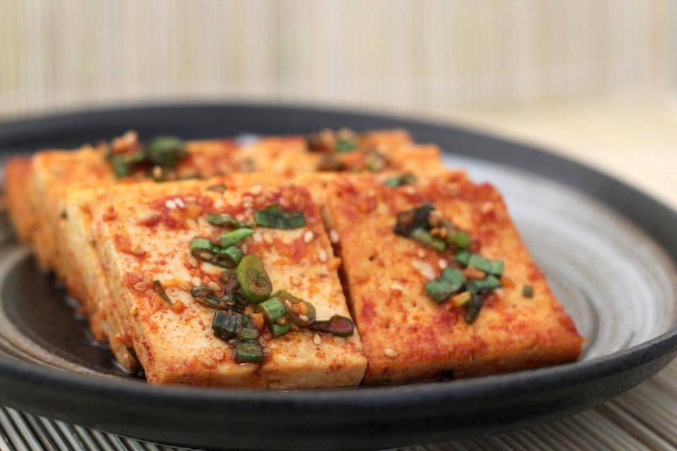 A plate of Sizzling Tofu, featuring golden-brown tofu slices topped with a flavorful sauce, garnished with chopped green onions and sesame seeds. The tofu has a crispy exterior and is seasoned to perfection, offering a delicious and spicy kick in every bite.