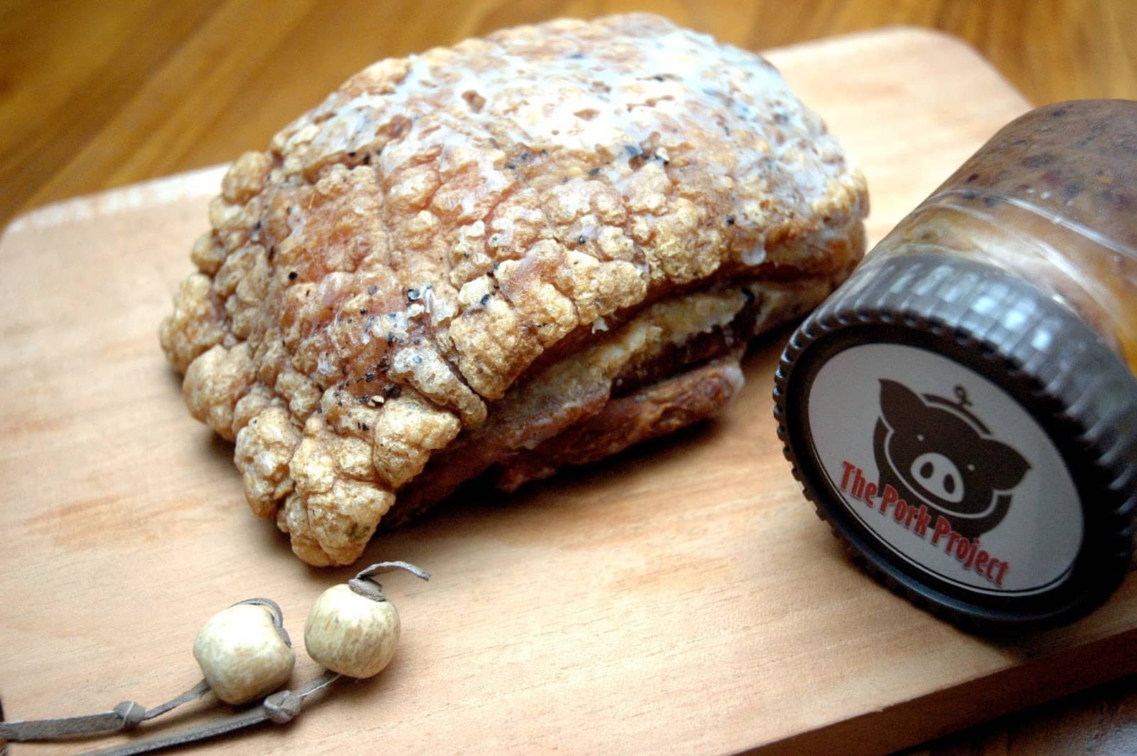 A perfectly crisp and golden-brown piece of bagnet (crispy pork belly) rests on a wooden cutting board, its crackling skin showcasing a delicious crunch. Beside the bagnet is a small jar labeled 'The Pork Project,' suggesting a sauce accompaniment.