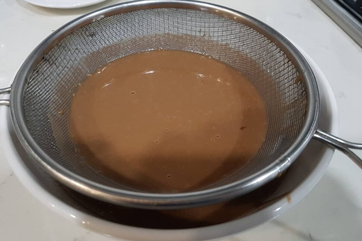 The image seems to depict a chocolate mixture being strained, which might be a step in preparing a dessert, possibly related to a dish like "Steam Milo Cheese." Milo is a popular chocolate malt powder often used in Filipino desserts.