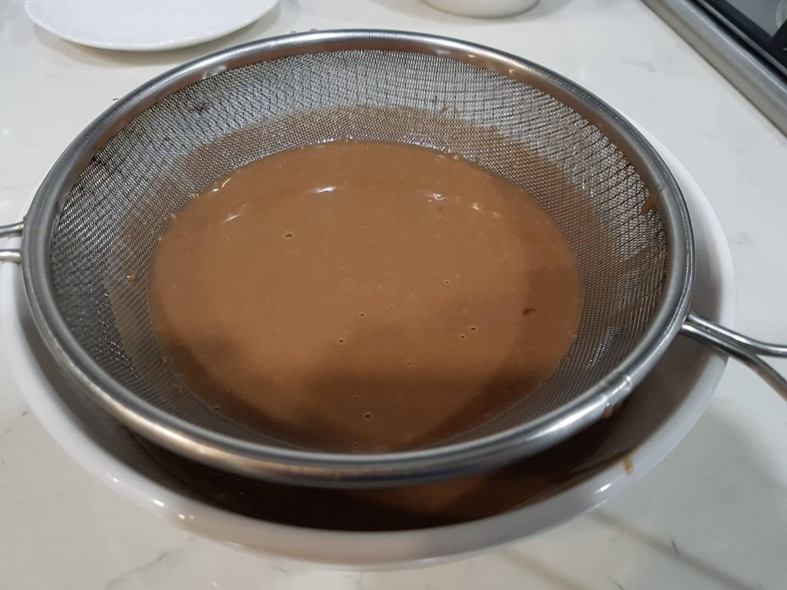 The image seems to depict a chocolate mixture being strained, which might be a step in preparing a dessert, possibly related to a dish like "Steam Milo Cheese." Milo is a popular chocolate malt powder often used in Filipino desserts.