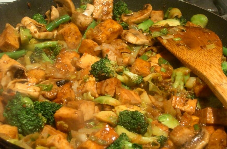 A sizzling pan of Stir Fry Kangkong and Pechay with Tofu, featuring golden-brown tofu cubes, fresh mushrooms, and vibrant green vegetables like broccoli and green beans. The dish is mixed with a savory sauce, giving it a glossy finish, while a wooden spatula rests on the side, ready to serve this delicious and healthy meal.