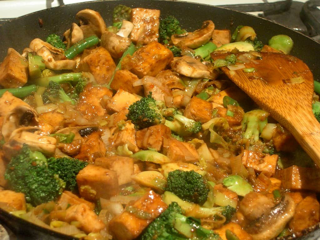 A sizzling pan of Stir Fry Kangkong and Pechay with Tofu, featuring golden-brown tofu cubes, fresh mushrooms, and vibrant green vegetables like broccoli and green beans. The dish is mixed with a savory sauce, giving it a glossy finish, while a wooden spatula rests on the side, ready to serve this delicious and healthy meal.