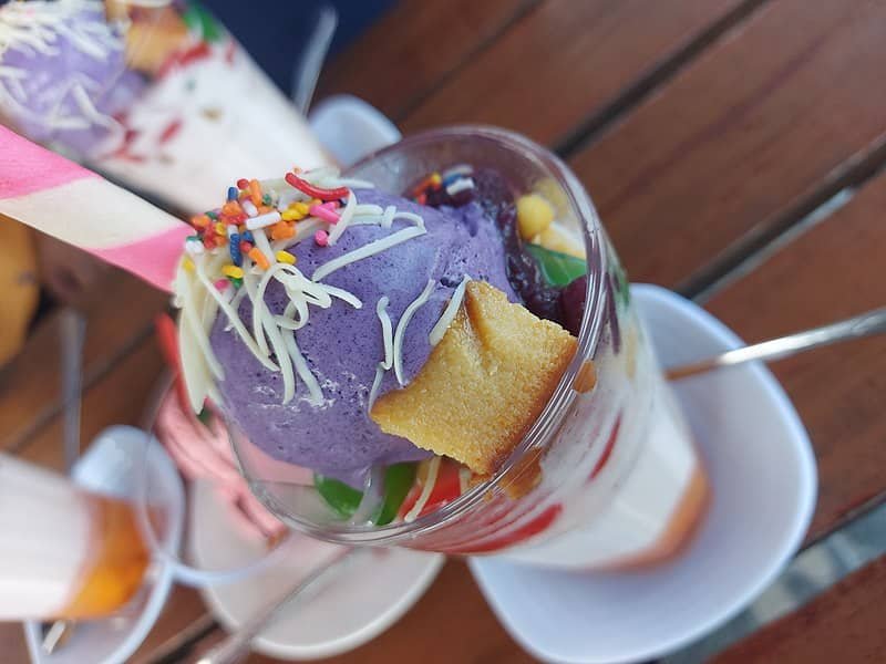 A colorful serving of halo-halo, a popular Filipino dessert, featuring ube ice cream, shaved ice, jelly, sweetened fruits, and leche flan, topped with sprinkles, cheese, and a wafer stick.