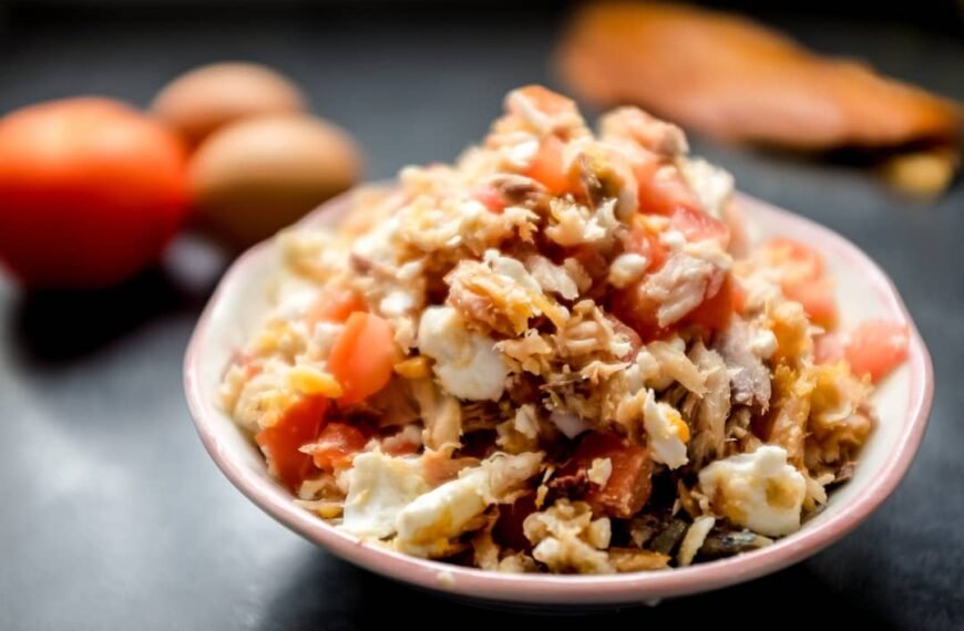 A close-up view of a dish of Tinapa Pasta, featuring flaked smoked fish (tinapa), diced tomatoes, and mixed vegetables served in a bowl. In the background, there are eggs and a fresh tomato, hinting at the ingredients used in the recipe.