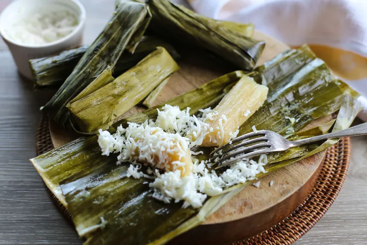 A traditional Filipino kakanin delicacy wrapped in banana leaves, unwrapped to reveal a soft, sticky rice treat topped with freshly grated coconut. The banana leaves, serving as a natural packaging, are laid open on a round wooden board, creating an inviting and authentic presentation. A fork rests beside the kakanin, ready for a delightful bite of this beloved Filipino snack.