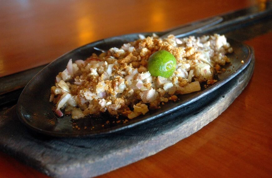 A sizzling plate of tuna sisig served on a hot iron skillet. The finely chopped tuna is mixed with onions, crunchy pork bits, and topped with a calamansi lime, ready to be squeezed for a tangy kick. The dish is placed on a rustic wooden board, enhancing its presentation.