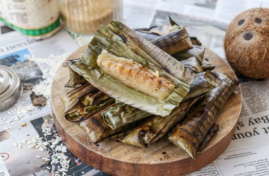 A serving of Tupig, a traditional Filipino delicacy made from glutinous rice and coconut, grilled in banana leaves. The banana leaf is peeled back, revealing the golden-brown, slightly charred exterior of the sticky rice dessert, placed on a wooden board with additional wrapped tupig in the background.