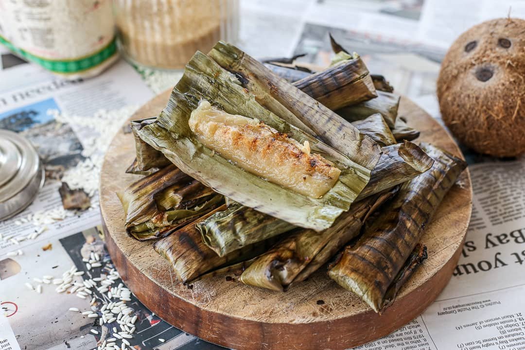 A serving of Tupig, a traditional Filipino delicacy made from glutinous rice and coconut, grilled in banana leaves. The banana leaf is peeled back, revealing the golden-brown, slightly charred exterior of the sticky rice dessert, placed on a wooden board with additional wrapped tupig in the background.