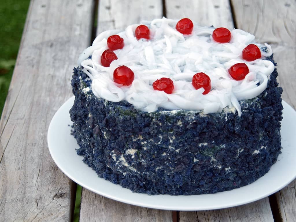 Ube Macapuno Roll Recipe - A rich purple ube cake covered with coconut strings and topped with red cherries, displayed on a white plate on a wooden table.