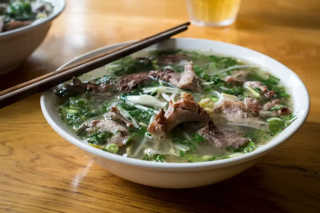 A steaming bowl of Vietnamese Pho Soup, filled with tender slices of beef, fresh herbs, and rice noodles in a clear, savory broth. The soup is garnished with chopped green onions and cilantro, and a pair of wooden chopsticks rests on the bowl's rim, ready to enjoy this traditional, aromatic dish.