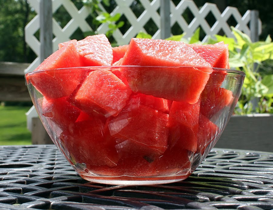 A clear glass bowl filled with large, juicy cubes of watermelon, set on an outdoor table with a lattice fence and garden plants in the background, under natural sunlight.