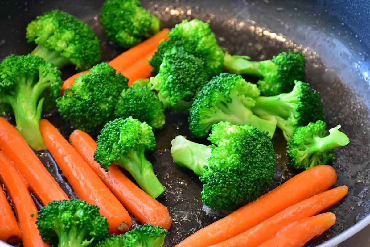 A vibrant bowl of buttered vegetables consisting of broccoli, carrots, corn, snow peas, and cauliflower. The vegetables are steamed to a perfect tenderness and lightly coated in butter, enhancing their natural flavors. They are presented in a white bowl set against a rustic blue-patterned tablecloth, emphasizing the freshness and colorful variety of the dish.