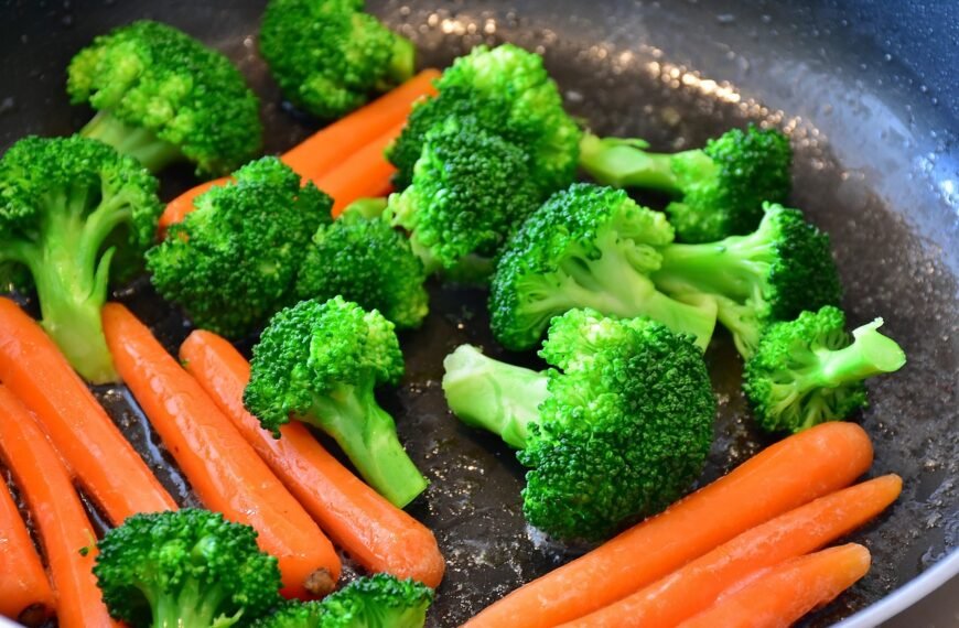 A vibrant bowl of buttered vegetables consisting of broccoli, carrots, corn, snow peas, and cauliflower. The vegetables are steamed to a perfect tenderness and lightly coated in butter, enhancing their natural flavors. They are presented in a white bowl set against a rustic blue-patterned tablecloth, emphasizing the freshness and colorful variety of the dish.