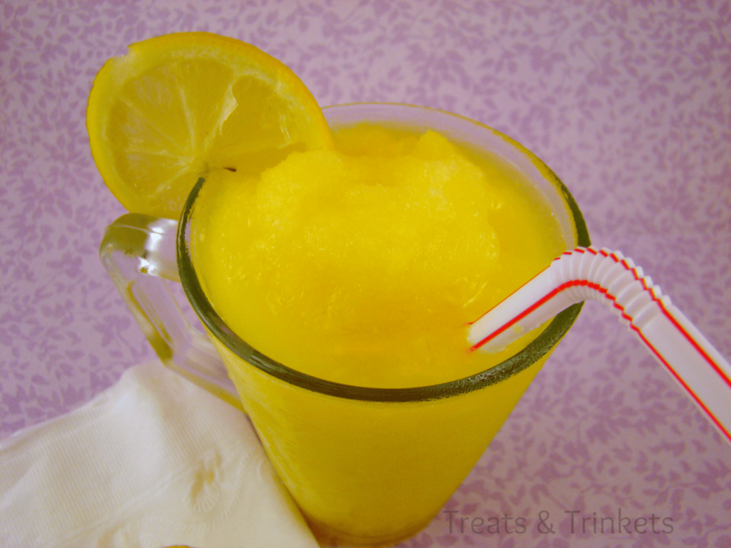 A vibrant yellow mango cooler served in a glass mug with a slice of lemon on the rim and a red-striped white straw, against a soft purple floral background.