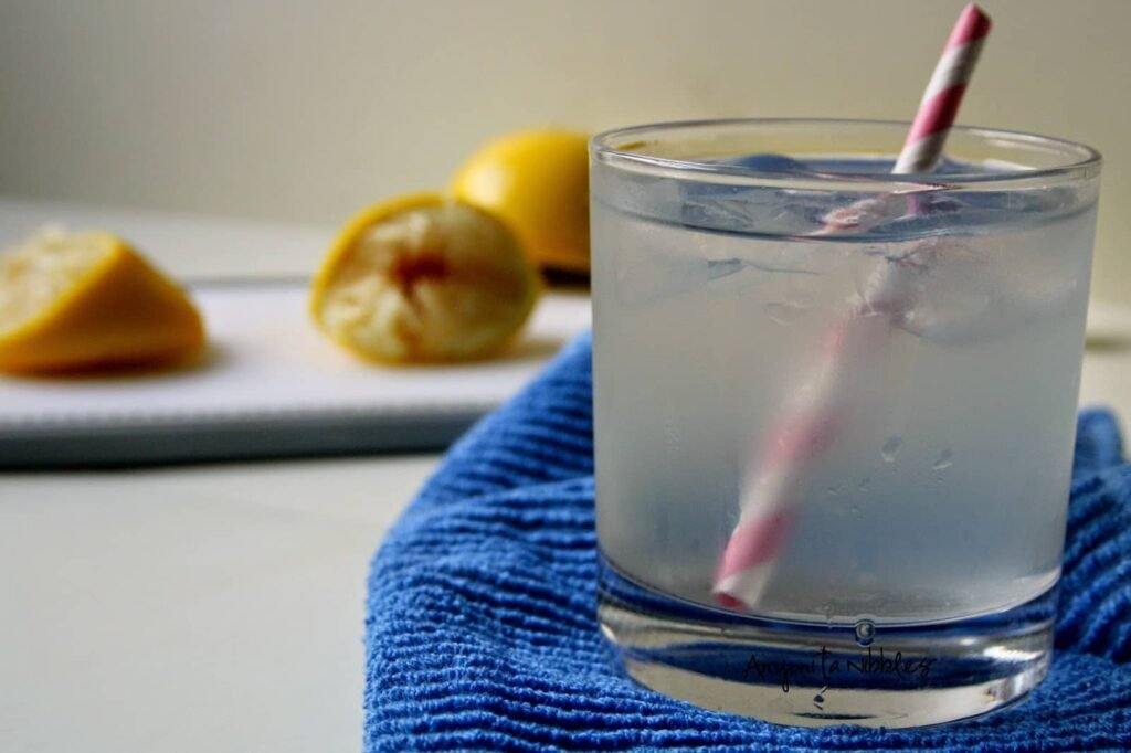 A refreshing glass of sparkling lemonade with ice, served with a striped pink straw on a blue towel, with freshly squeezed lemons in the background on a white cutting board.